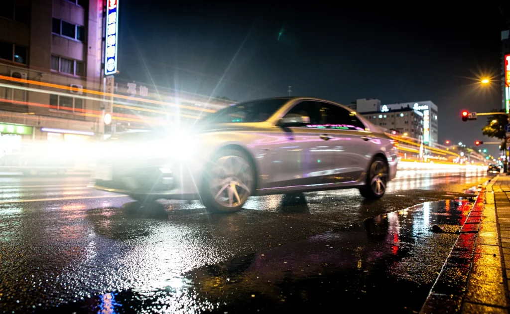 Voiture en mouvement rapide photographiée la nuit par un radar, phares allumés, avec des reflets de lumières urbaines sur l’asphalte mouillé.