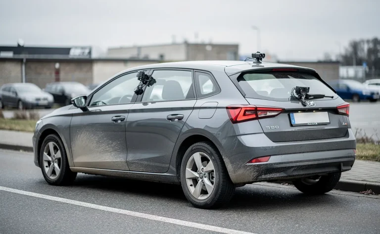 Une voiture grise type hatchback, discrètement garée au bord de la route, présente des caméras et capteurs sur le tableau de bord et la lunette arrière.