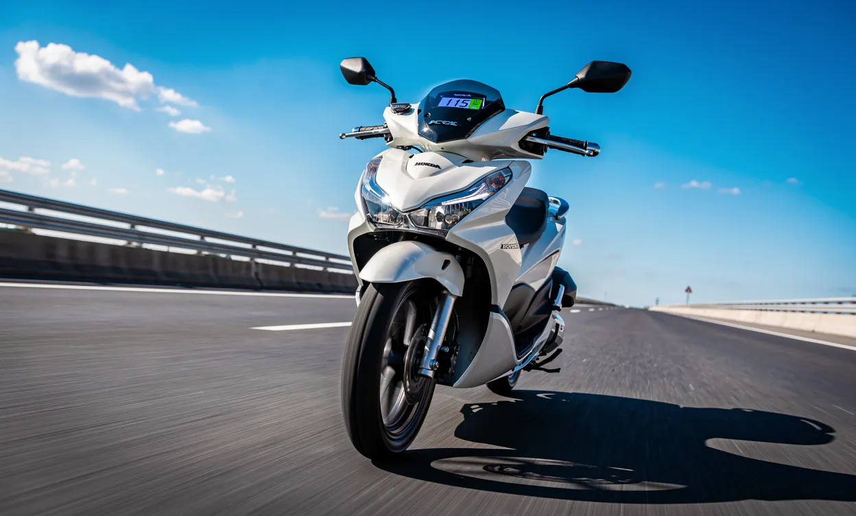 Un scooter Honda PCX 125 roule à 115 km/h sur une route dégagée sous un ciel bleu avec des détails chromés brillants et une perspective dynamique en contre-plongée.